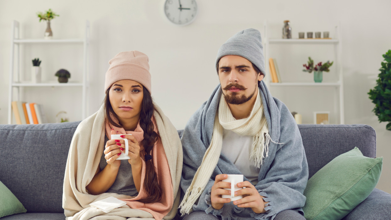 Dissatisfied young couple having problem with central heating, sitting on sofa at home, freezing, drinking hot tea trying to warm up. Sick man and woman wrapped in blankets suffering from cold or flu