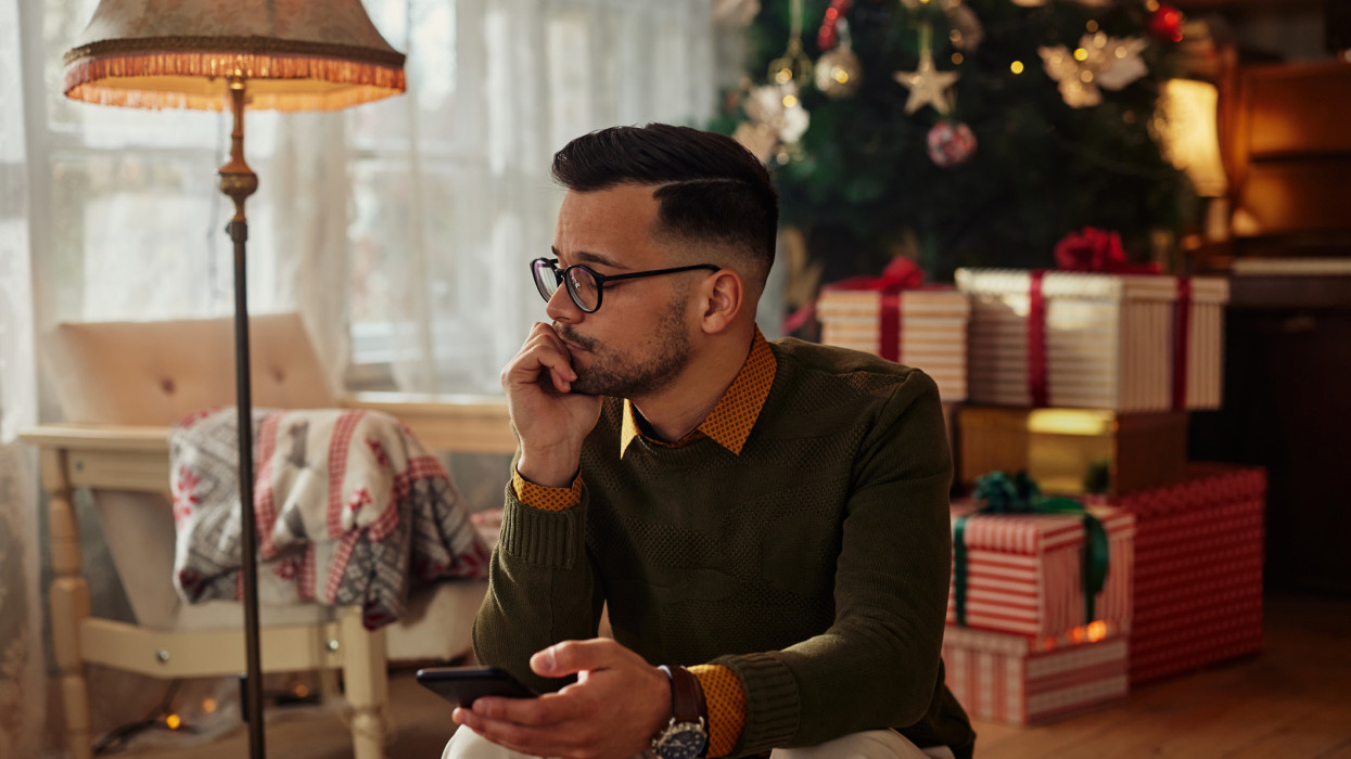 Sad thoughtful Xmas man typing text message on smartphone, chatting online, sitting beside Christmas tree. Man with mobile phone feeling sad, frustrated, lonely at Christmastime